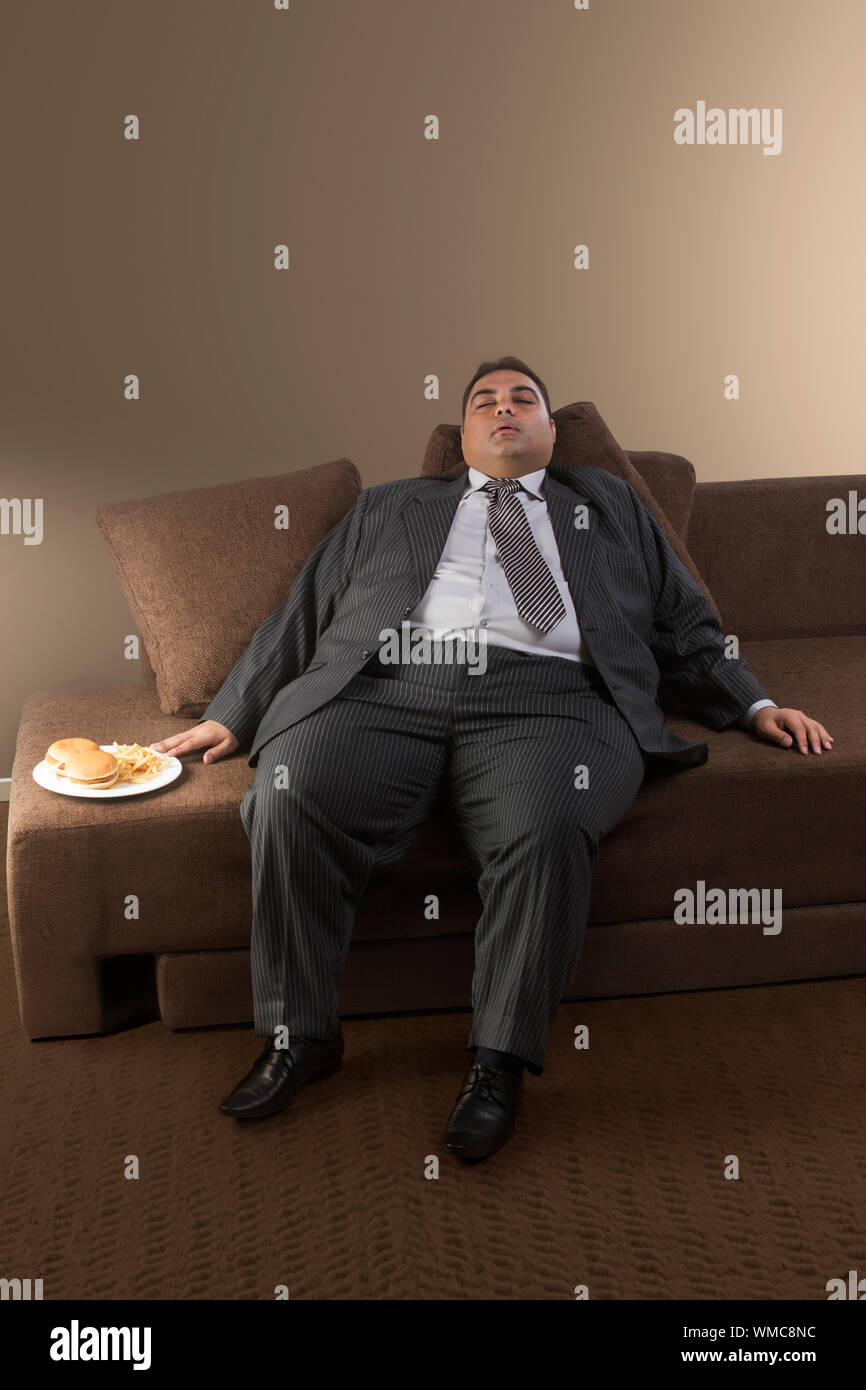 Homme d'obèses dans les vêtements officiels de dormir sur un canapé avec la plaque d'hamburgers et de frites à ses côtés Banque D'Images