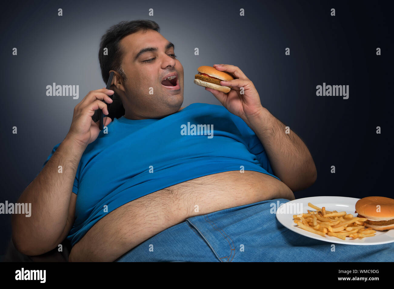 L'homme obèse détendue avec ventre visible hors de sa chemise de manger tout en parlant plus de burger téléphone mobile avec une assiette de hamburger et frites sur son thi Banque D'Images