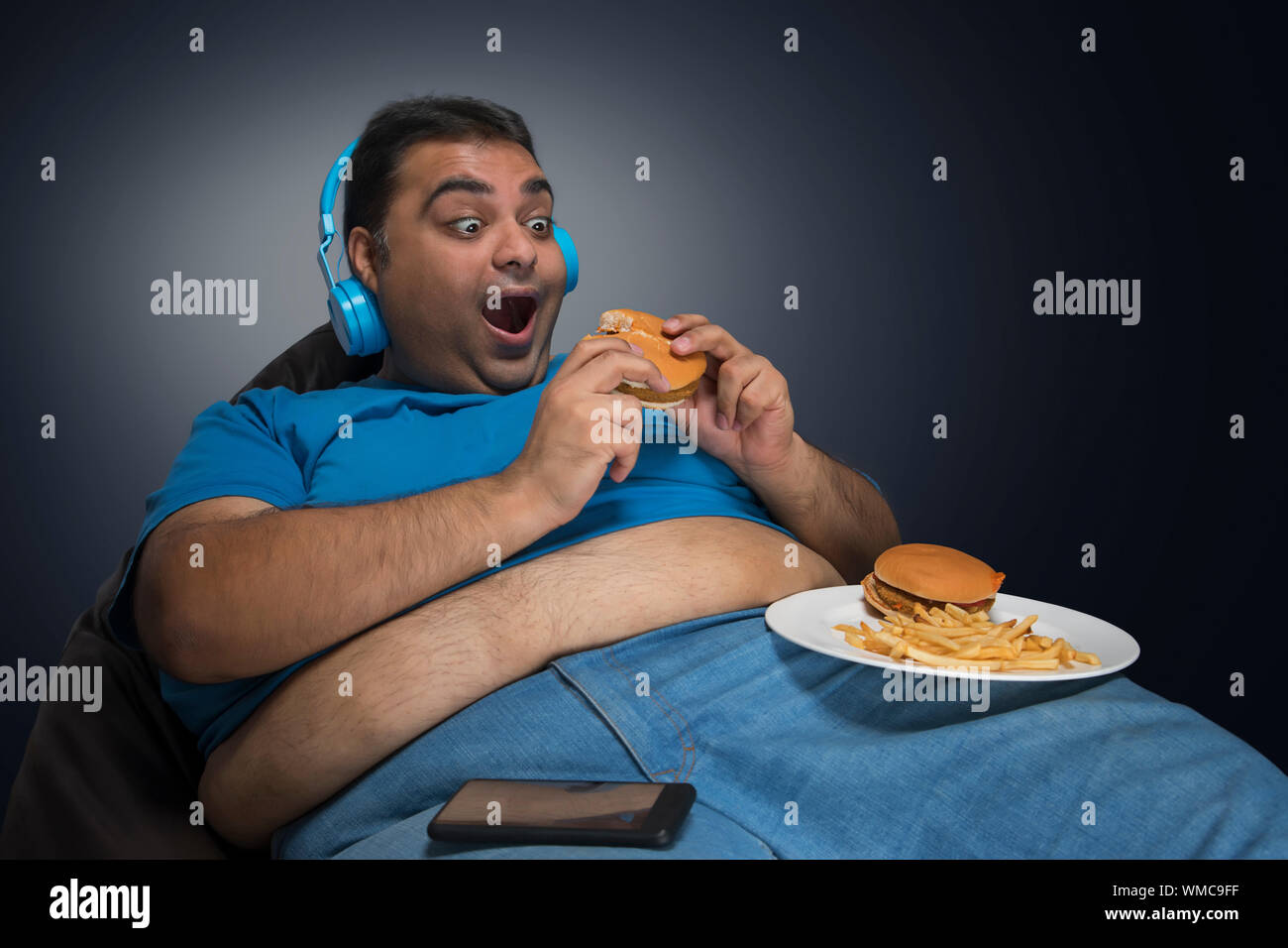 L'homme obèse excité avec ventre visible hors de sa chemise manger burger tout en écoutant de la musique avec une assiette de hamburger et frites sur sa cuisse Banque D'Images
