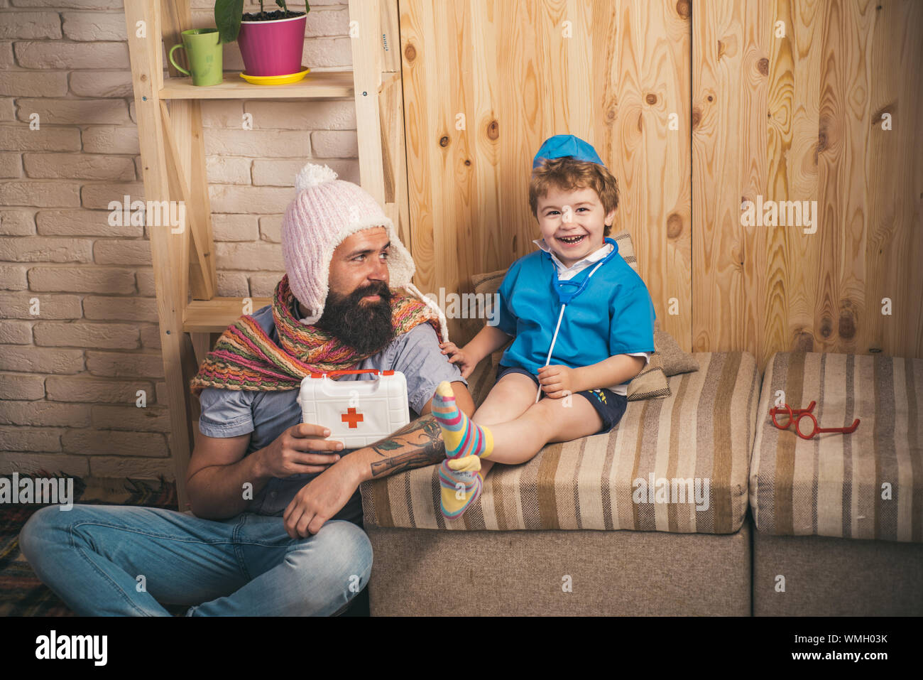 Jeu de la famille l'hôpital, les soins de concept. Père comme patient et kid comme médecin. Papa avec visage souriant, longue barbe et moustache, élégant et gai Banque D'Images