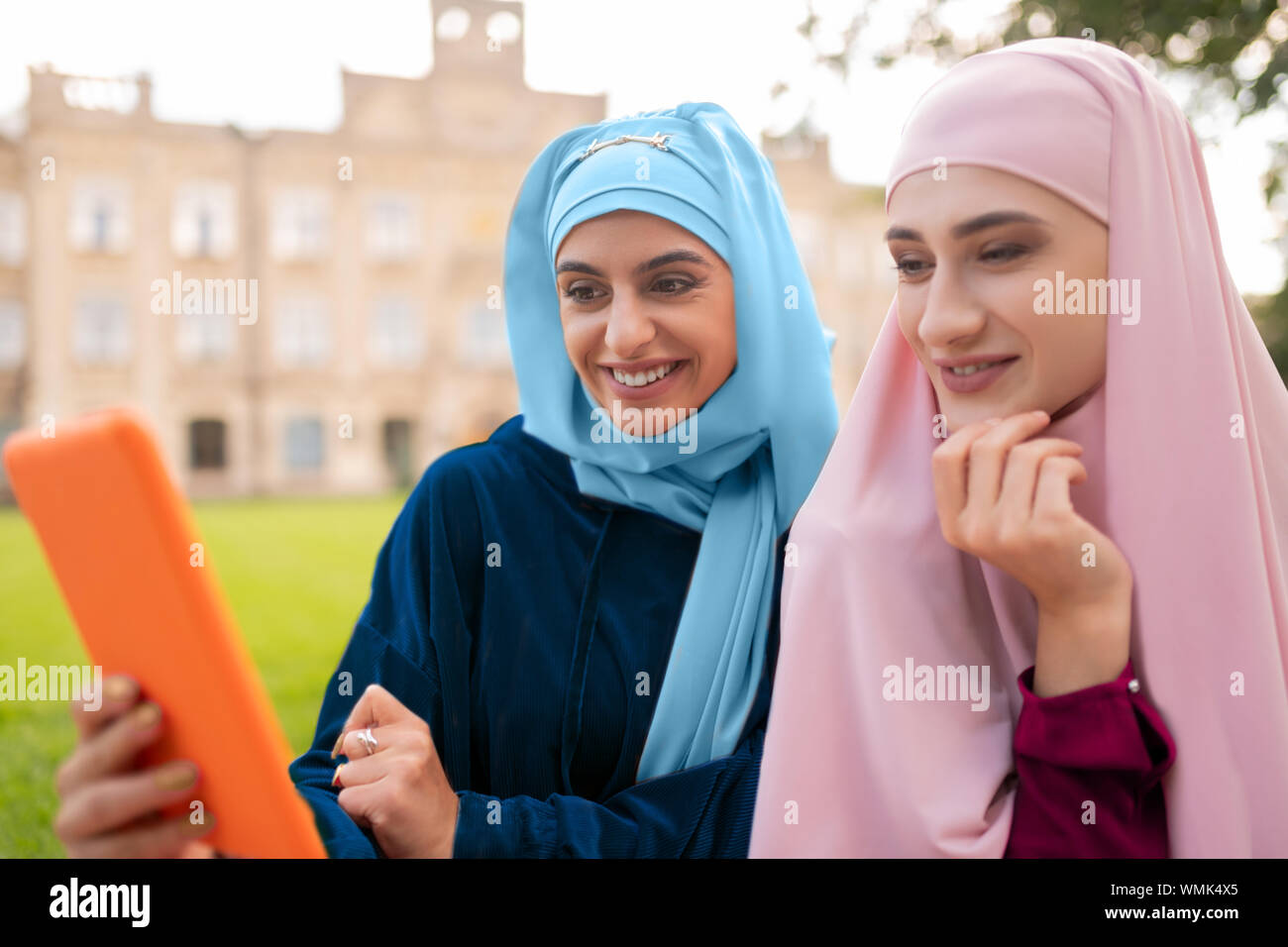 Student wearing hijab bleu holding orange tablet assis près de ami Banque D'Images