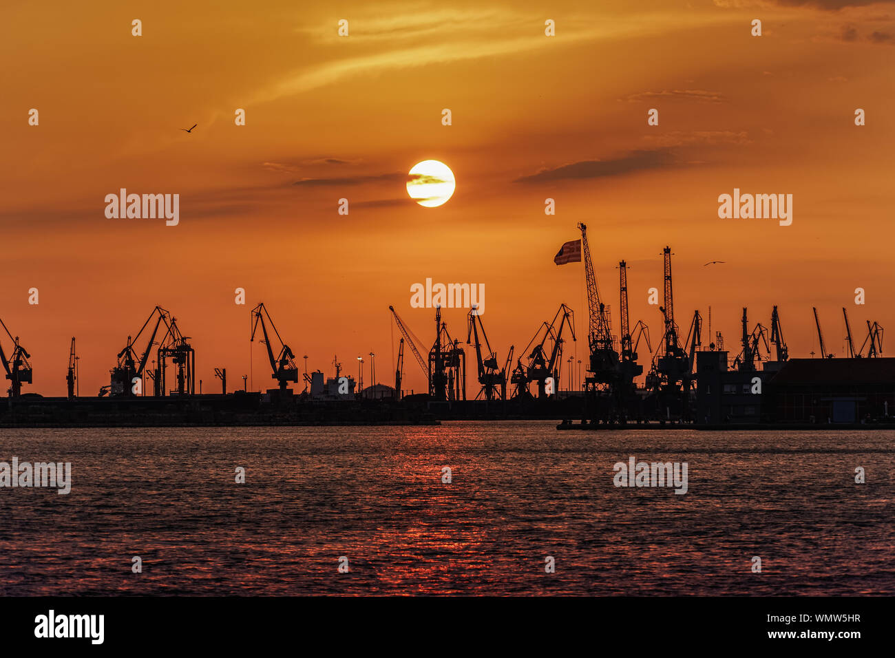 Thessalonique, Grèce heure golden sunset paysage à la ville port. Coucher du soleil orange au-dessus du port de faible hauteur des bâtiments, avec des grues portuaires et de brandir le drapeau grec. Banque D'Images