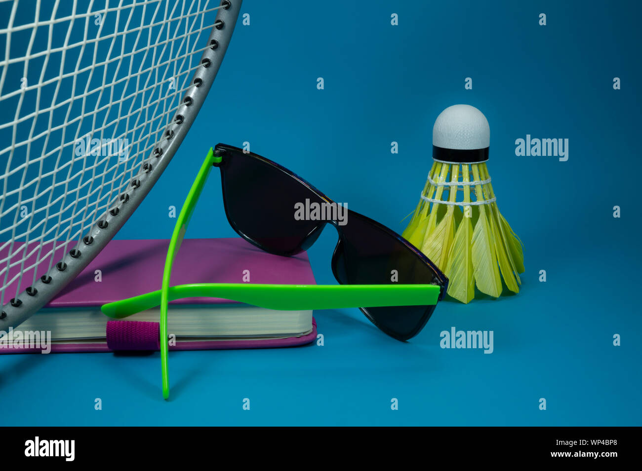 Volants de badminton raquette colorés avec des lunettes de soleil et se reposer sur un livre rose de couleur vive ou de journal sur un fond bleu dans un close up encore Banque D'Images