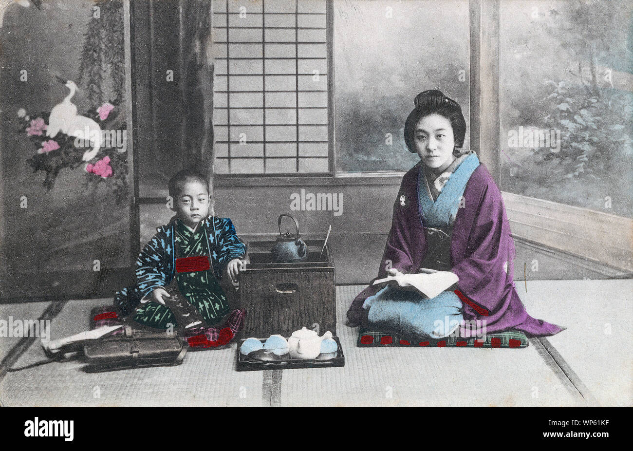 [ 1900 - Japon Japanese Woman and Boy en Kimono ] - une jeune femme en kimono traditionnel et style de coiffure et un jeune garçon assis à côté d'un hakohibachi, un brasero enfermé dans une boîte en bois. La femme tient un livre. En face de la boîte se trouve un plateau avec une Théière et tasses à thé. Devant le garçon est un sac en cuir. 20e siècle vintage carte postale. Banque D'Images