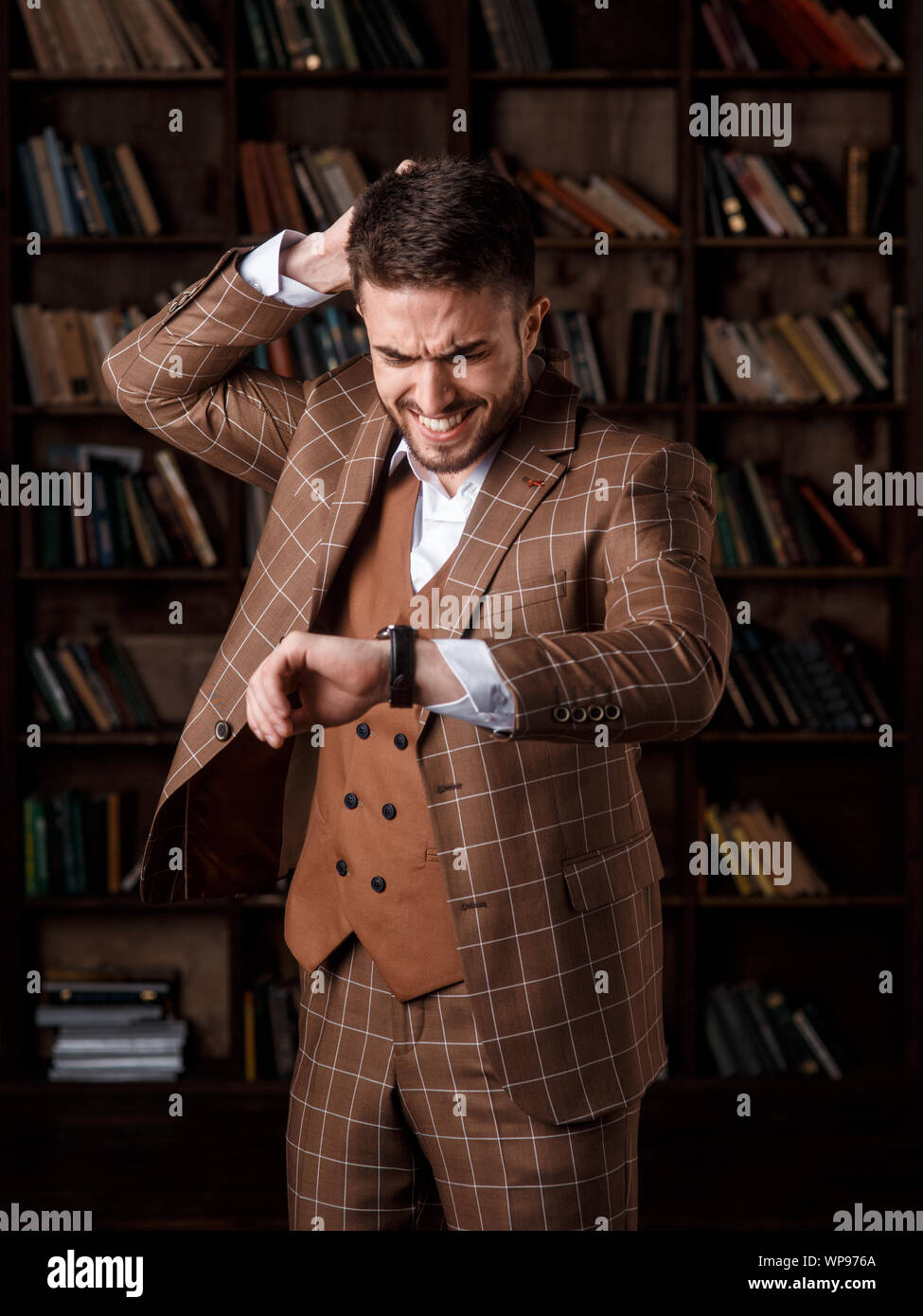 Un jeune homme avec une barbe dans un costume brun avec un sourire se repose sur sa montre et tient sa tête tout en se tenant dans la bibliothèque. Un homme sourit et assure le suivi des temps Banque D'Images