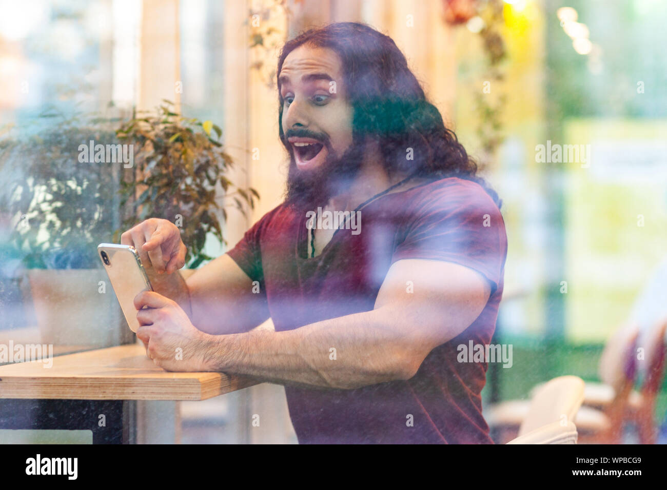 Portrait vue latérale par la fenêtre d'excité beau jeune homme sportif adultes avec de longs cheveux bouclés assis sur cafe, holding phone avec surpris fa Banque D'Images