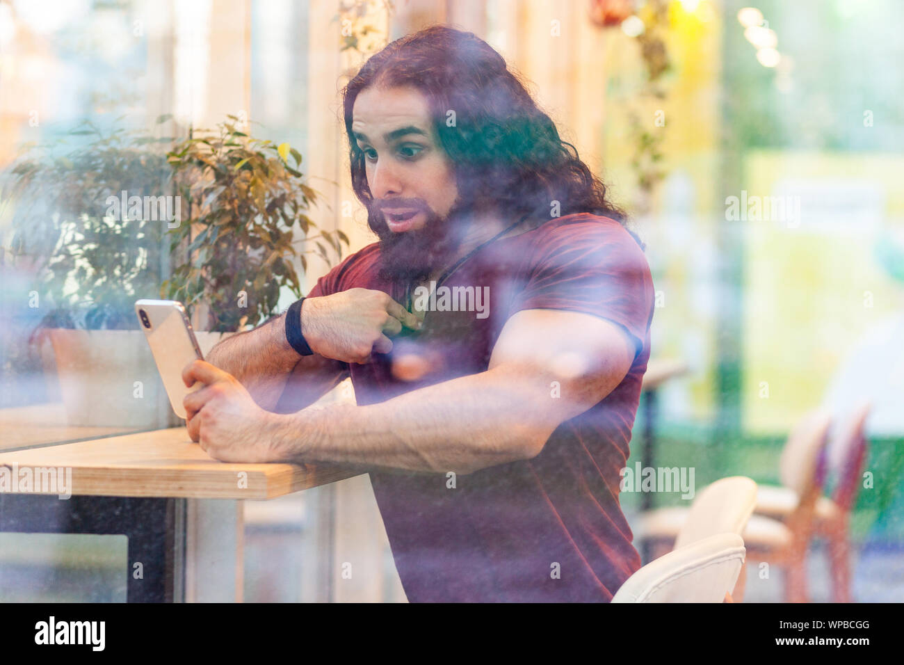 Portrait à travers la fenêtre de l'incroyable beau jeune homme musclé adultes avec de longs cheveux bouclés assis sur cafe, holding phone avec choqué face po Banque D'Images