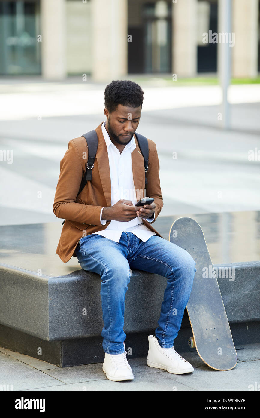 Portrait de jeune homme afro-américain de taper un message texte via smartphone tout en étant assis à l'extérieur en milieu urbain, copy space Banque D'Images