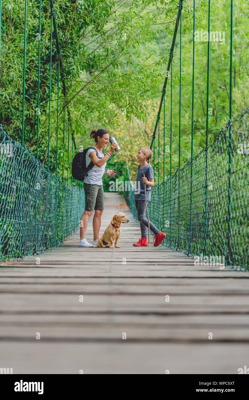 Mère et fille marcher sur le pont suspendu en bois avec petit chien jaune dans la forêt Banque D'Images Mère et fille marcher sur le pont suspendu en bois avec petit chien jaune dans la forêt Banque D'Images
