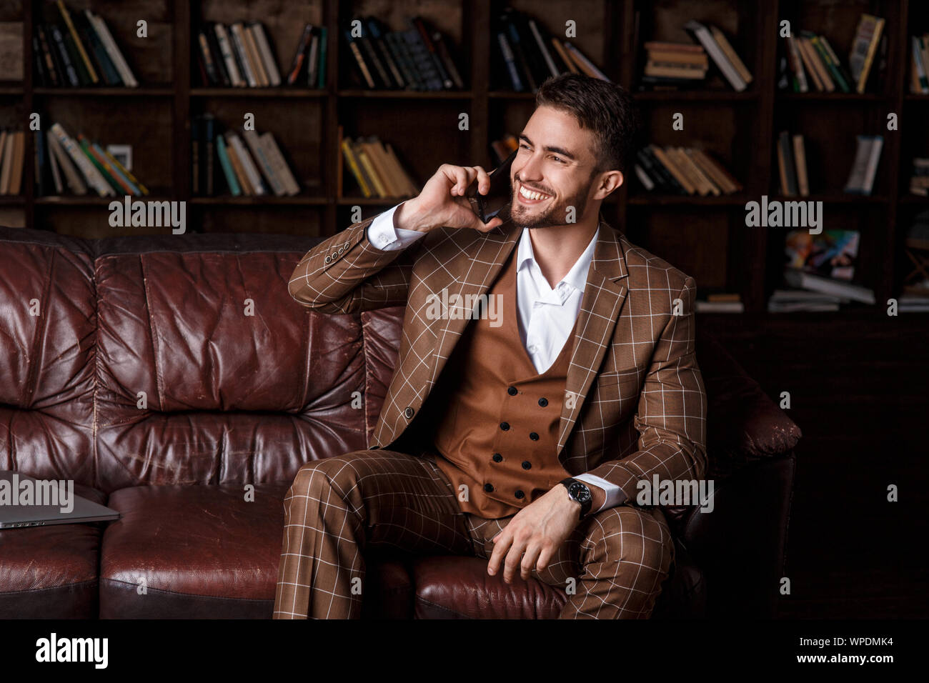 Jeune homme barbu dans un costume marron à parler au téléphone avec un sourire sur son visage alors qu'il était assis dans le bureau de la bibliothèque. Banque D'Images
