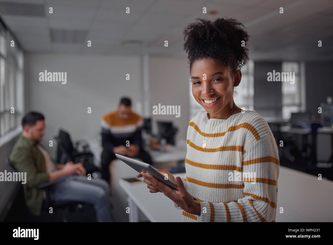 Cheerful young African American Woman in casual wear holding digital tablet souriant et à la recherche d'appareil alors que ses collègues de la communication Banque D'Images