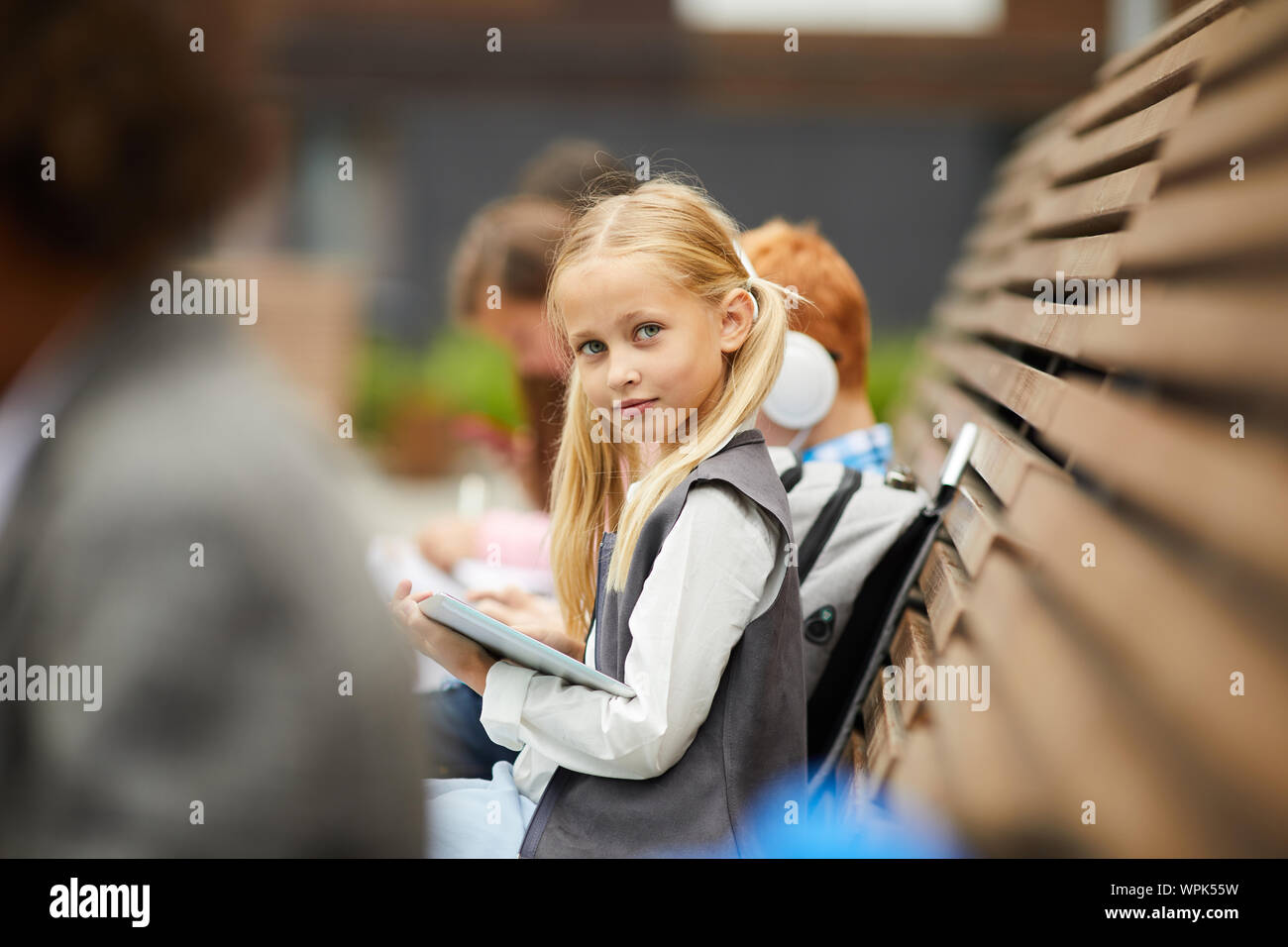 Portrait d'écolière avec de longs cheveux blonds looking at camera, assis sur le banc avec ses camarades et using digital tablet outdoors Banque D'Images