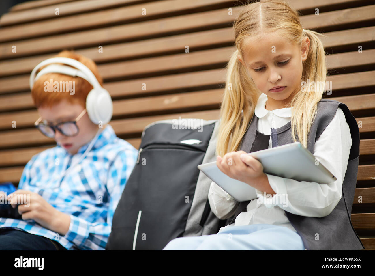 Cute schoolgirl avec de longs cheveux blonds using digital tablet, pour son étude, assise sur le banc d'écolier avec en arrière-plan Banque D'Images