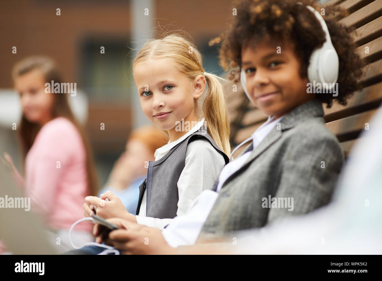 Portrait of cute lycéenne aux cheveux blonds looking at camera, assis sur le banc avec son camarade qui l'aide de mobile phone Banque D'Images
