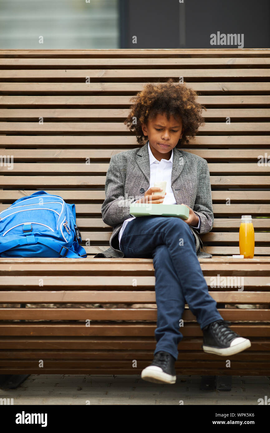 African boy eating sandwich et boire du jus tout en restant assis sur le banc à l'extérieur pendant les pauses à l'école Banque D'Images