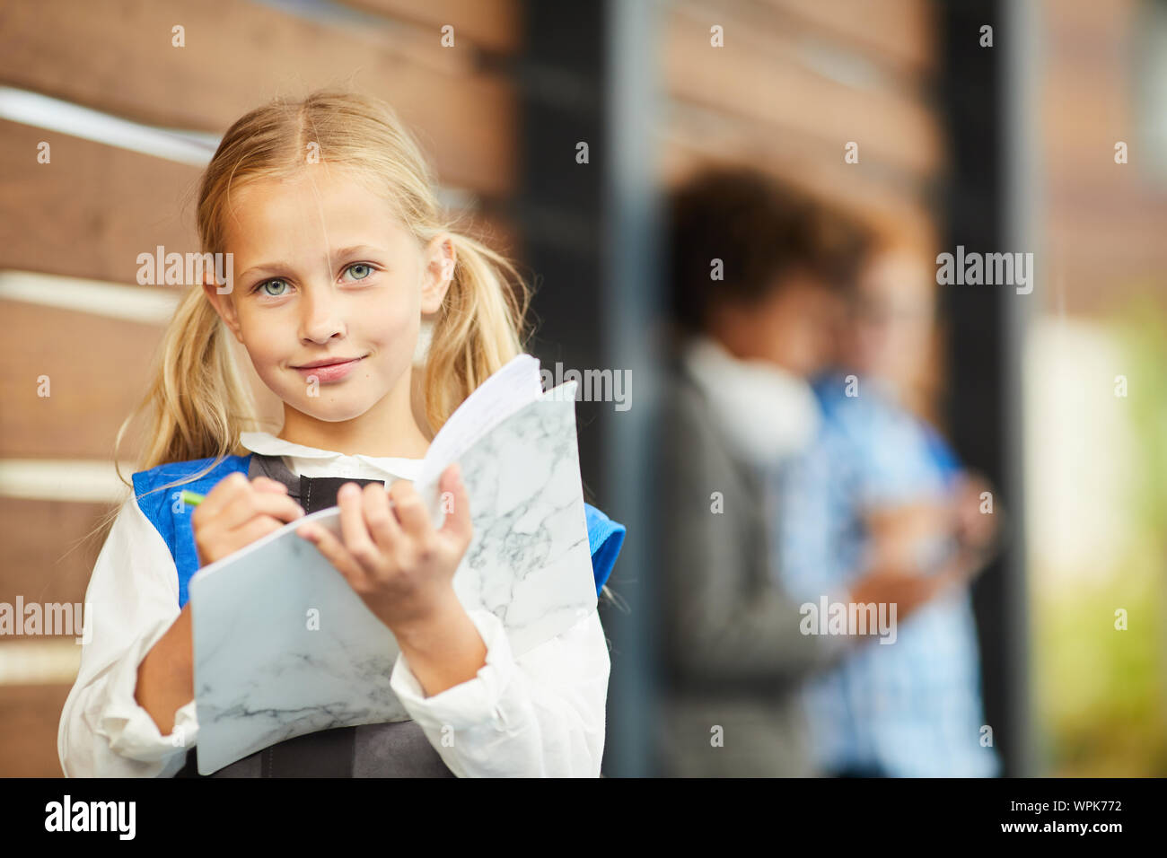 Portrait de petite fille aux longs cheveux blonds looking at camera, tout en faisant des notes en plein air pour manuels Banque D'Images