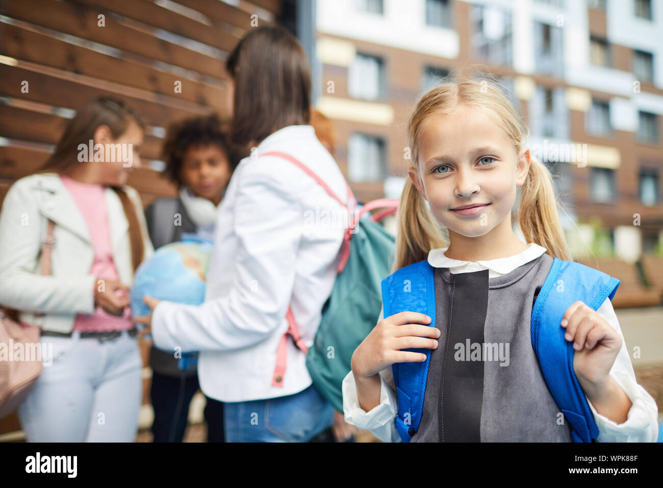 Portrait de petite écolière avec de longs cheveux blonds avec sac à dos derrière son dos smiling at camera avec d'autres enfants de l'école dans le fond pis Banque D'Images