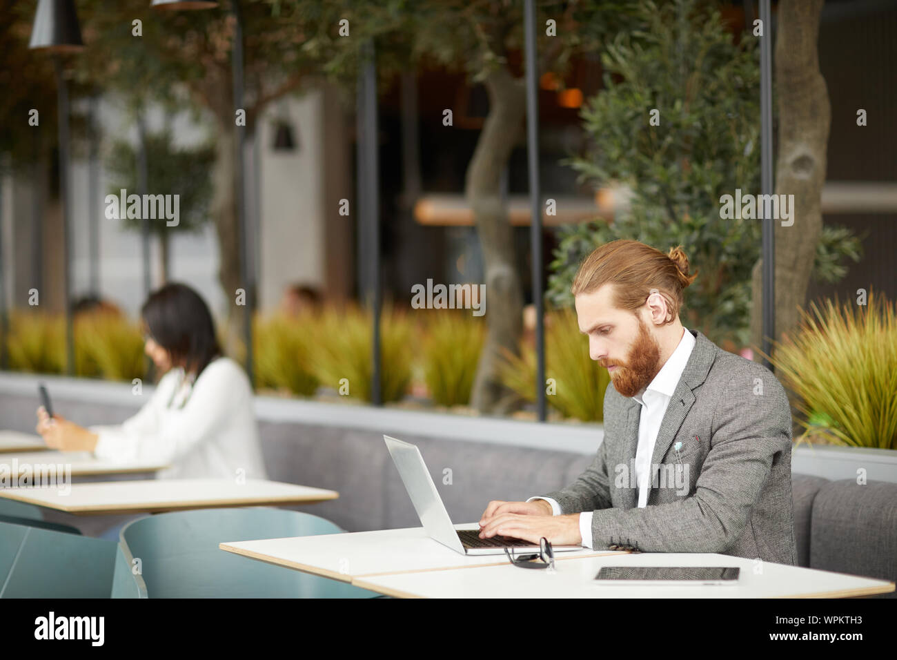 Jeune homme d'affaires graves en fonction de se concentrer sur son travail en ligne il la saisie sur ordinateur portable avec businesswoman en arrière-plan de cafe Banque D'Images