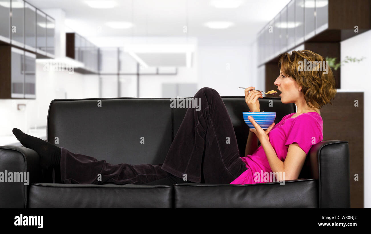 Les femmes de race blanche avec les cheveux crépus assise sur un sofa de manger un bol de céréales de petit-déjeuner. Elle est à la maison et regarde comme elle vient de se réveiller et de faire un mo Banque D'Images Les femmes de race blanche avec les cheveux crépus assise sur un sofa de manger un bol de céréales de petit-déjeuner. Elle est à la maison et regarde comme elle vient de se réveiller et de faire un mo Banque D'Images