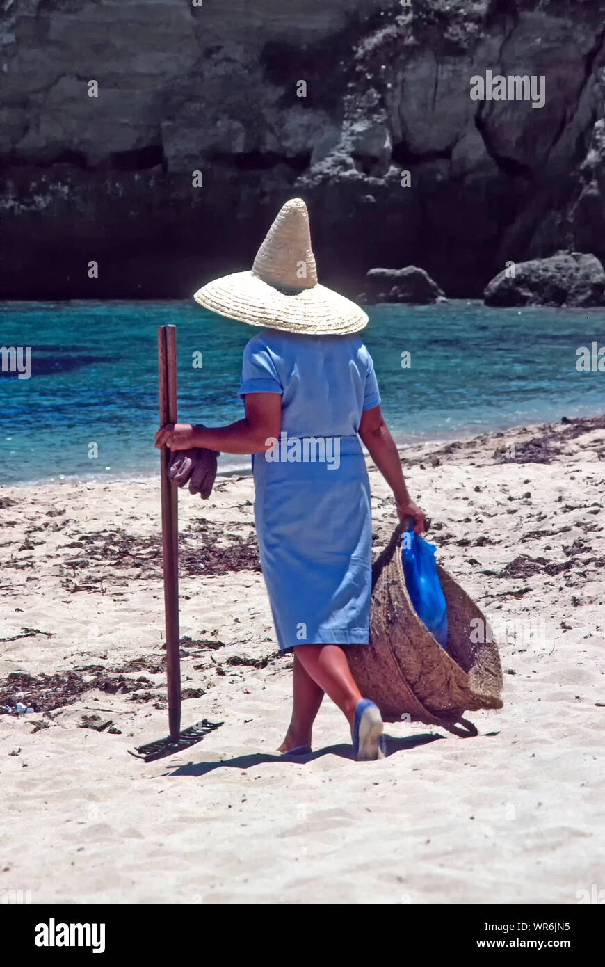 Femme marche pour travailler sur plage de sable fin 1983 Réalisation & râteau grand panier algues collecte portant chapeau de paille large bord fait chaud soleil Portimao algarve Banque D'Images