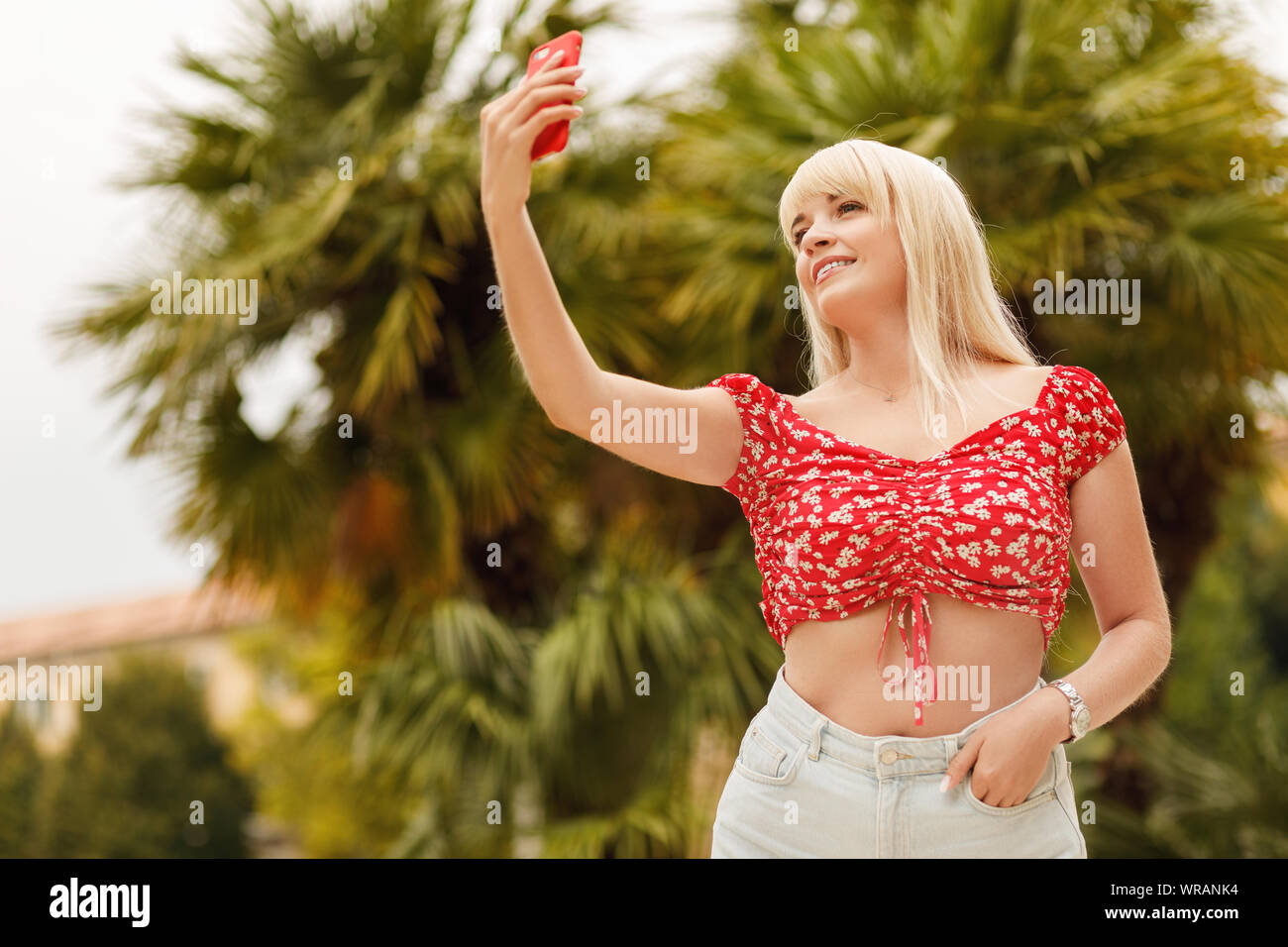 Jeune fille blonde gaie souriante debout sur fond de la rue des plantes tropicales et l'aide de smart phone. Banque D'Images