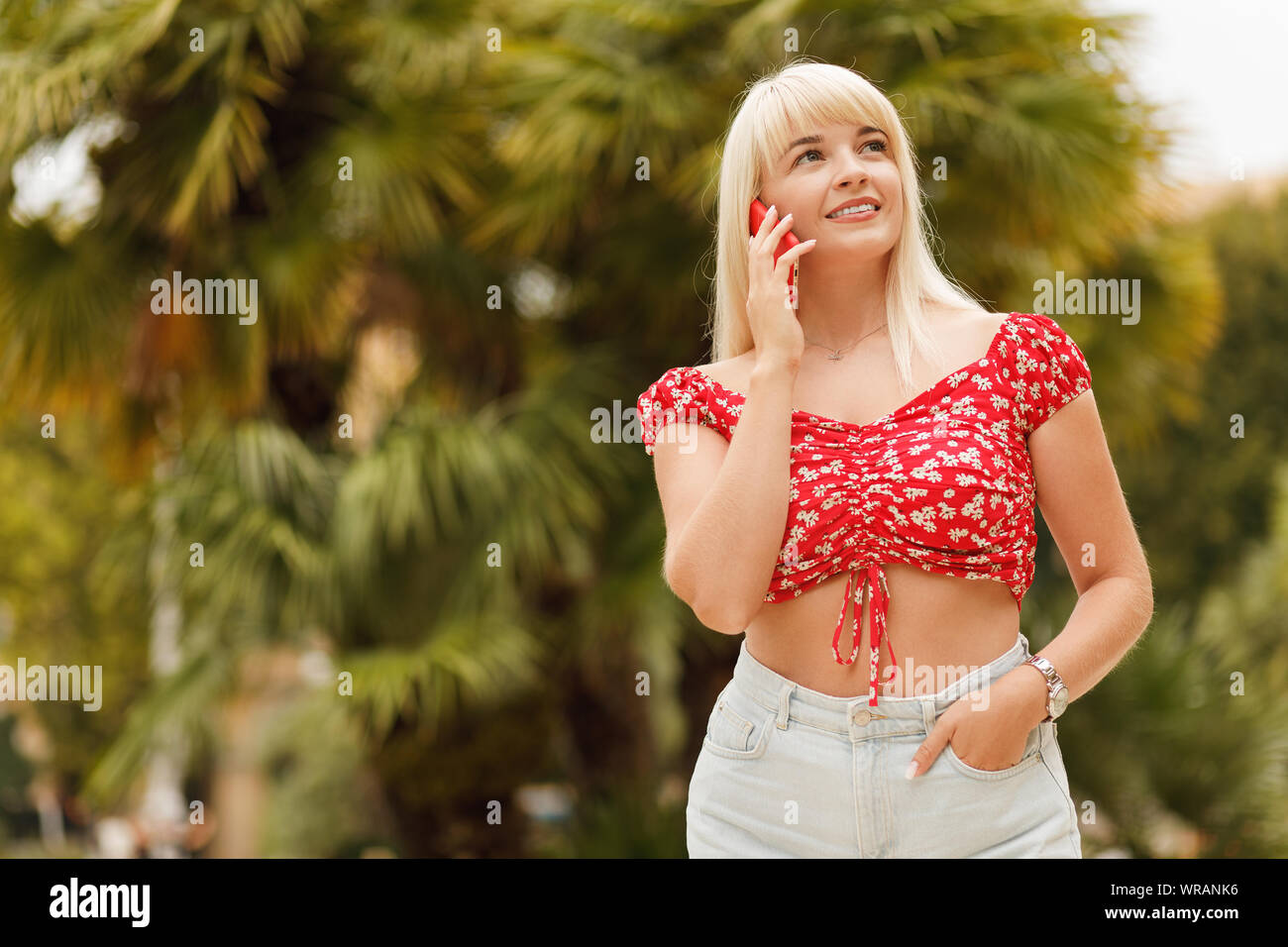 Jeune fille blonde gaie souriante debout sur fond de la rue des plantes tropicales et l'aide de smart phone. Banque D'Images