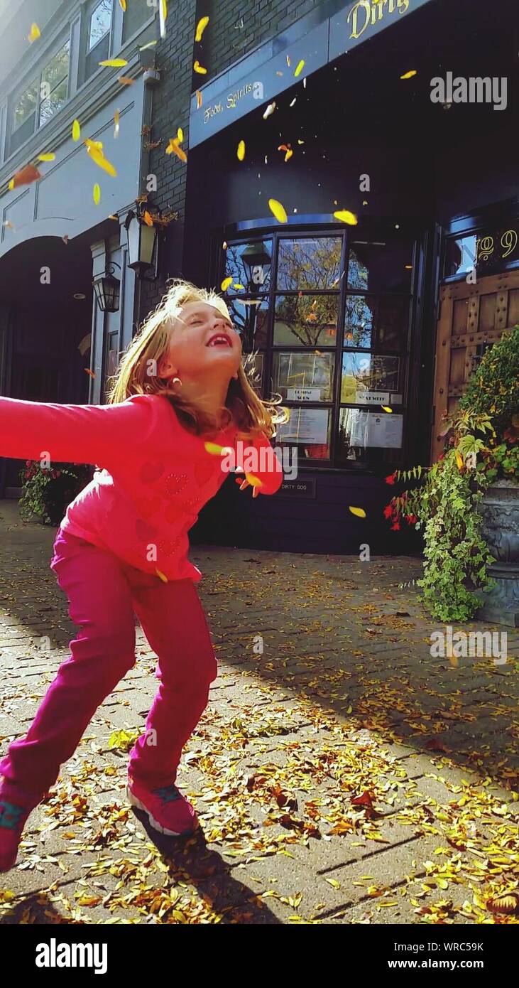 Jolie fille jouant avec les feuilles qui tombent sur le trottoir par des bâtiments en ville Banque D'Images