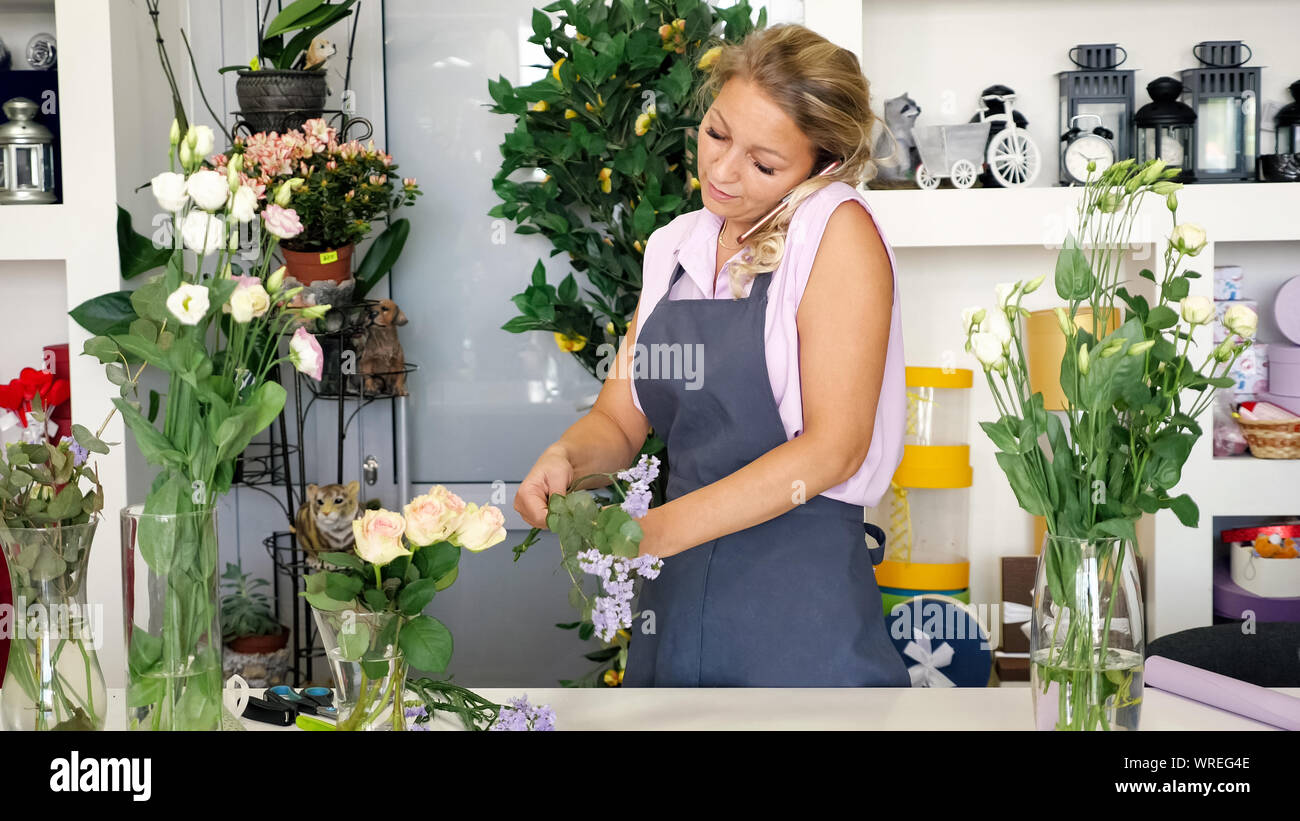 Fleuriste professionnel femme parle sur smartphone en prenant l'ordre et s'bouquet travaillant dans le magasin de fleurs. Mature Woman in apron dans art floral studio créer de belles bunch. Les personnes travaillant à leur concept. Banque D'Images