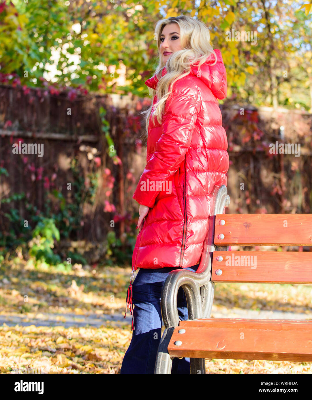 Girl porter du rouge clair veste chaude. Concept de la mode d'automne.  Belle Dame posant en veste près de banc. Femme blonde à la mode d'automne,  stand maquillage avec parc. Veste pour