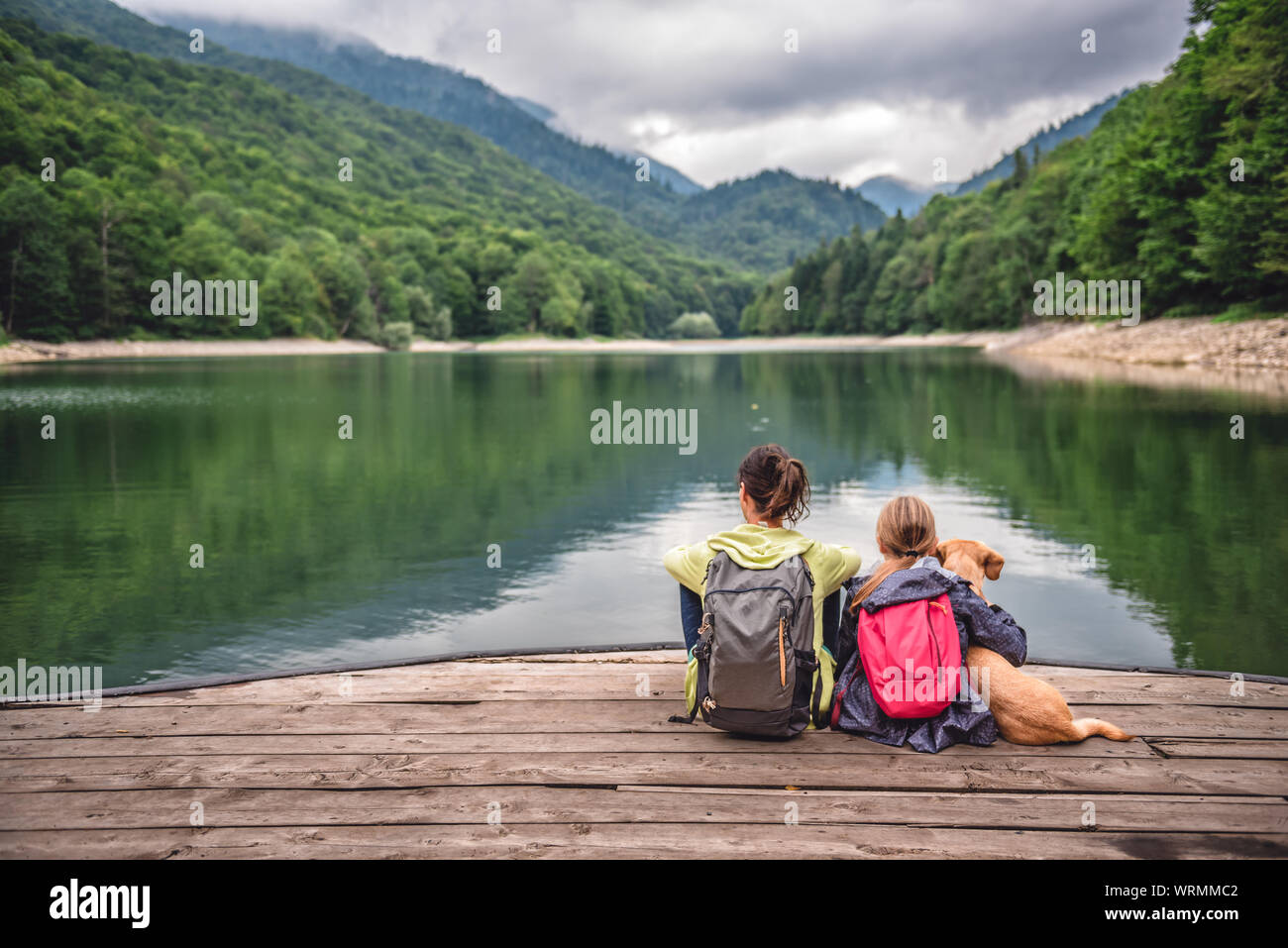 Mère et fille avec un petit chien jaune reposant sur un quai et à la recherche au lac et montagnes brumeux Banque D'Images