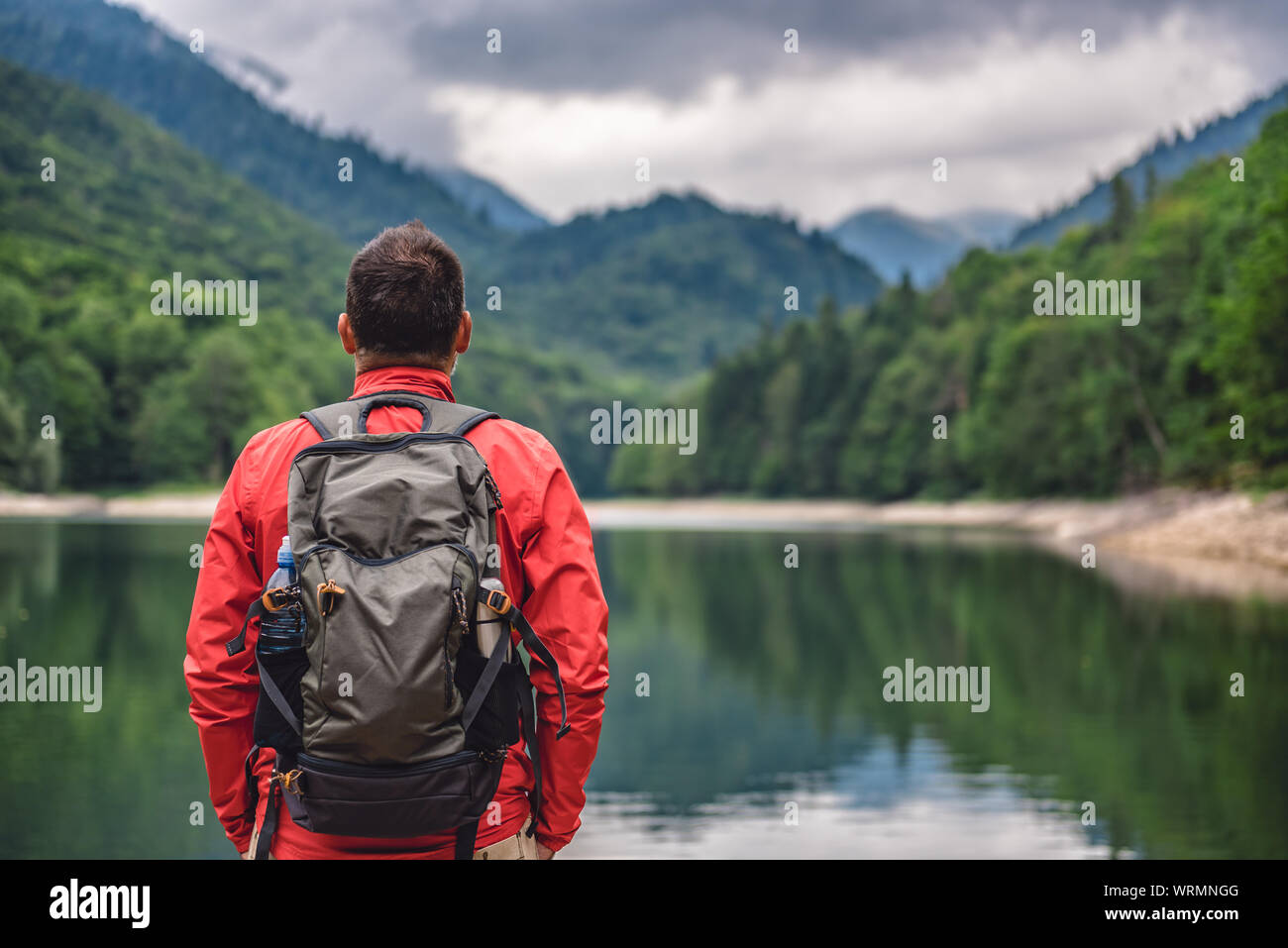 Randonneur wearing red jacket sac à dos et debout près du lac de montagne et à la recherche à la distance Banque D'Images