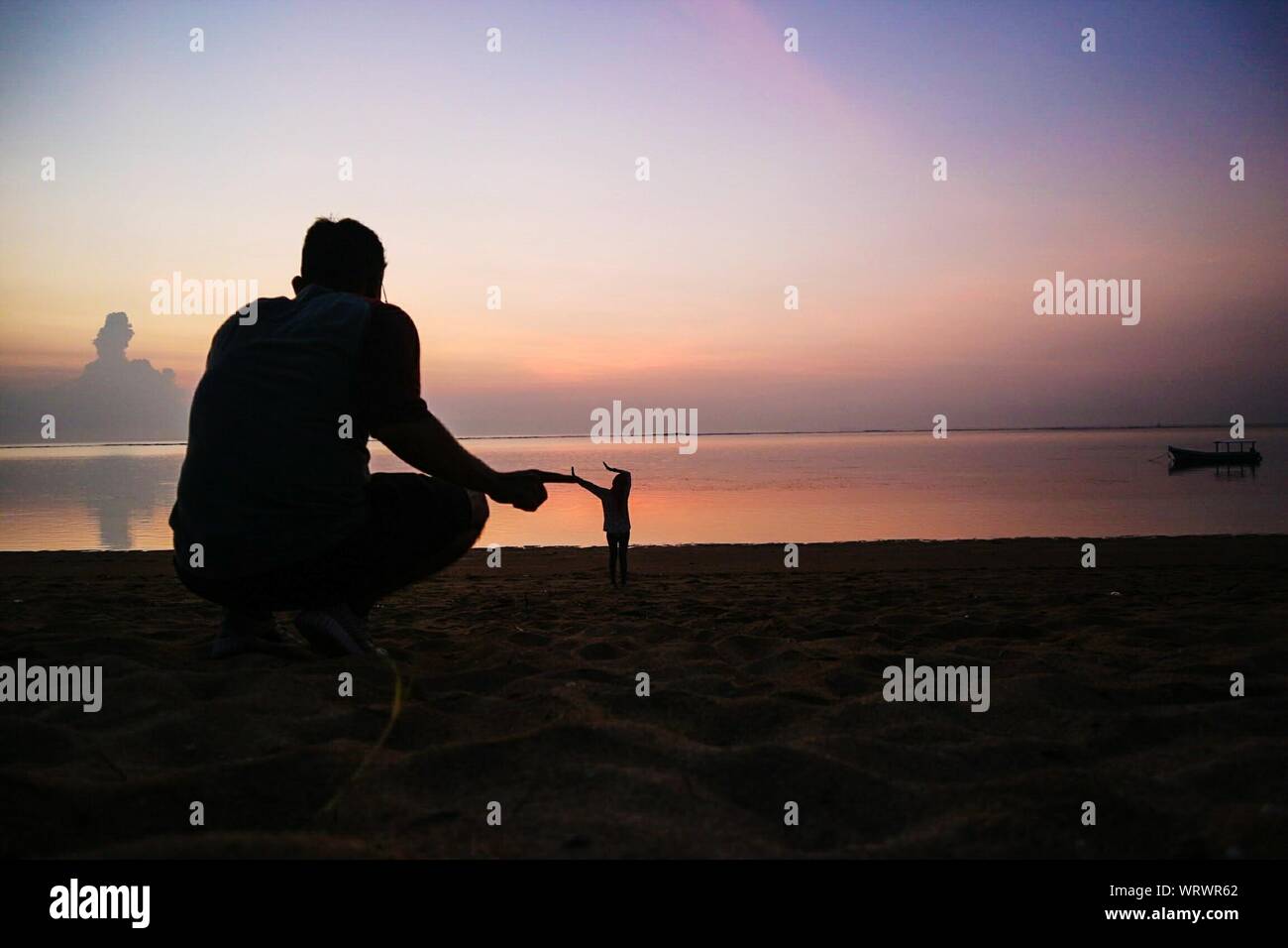 Illusion optique d'homme géant de toucher toute petite femme gesticulant en se tenant sur le rivage à la plage pendant le coucher du soleil Banque D'Images