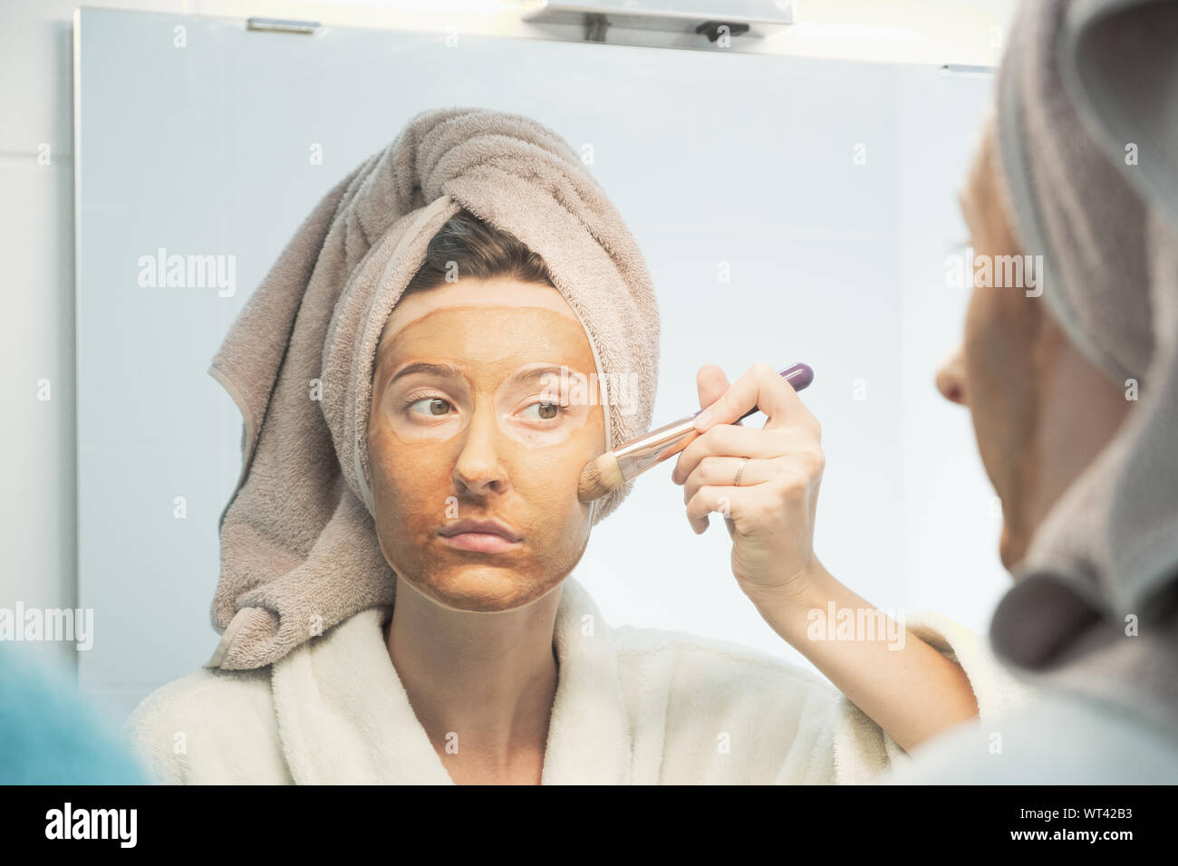 Jeune femme met sur son visage masque à l'argile à la brosse avec miroir de maquillage dans la salle de bains. Le concept de l'autogestion, bon soins du visage, soins quotidiens de la peau Banque D'Images