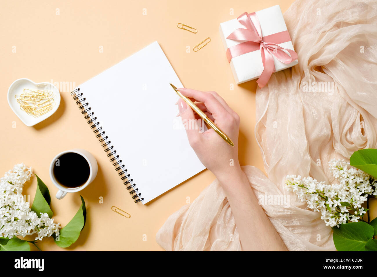 Haut de la vue, télévision laïques 24 avec des fleurs lilas, soie, tasse à café, boîte-cadeau, human hand holding golden pen et l'écriture dans le bloc-notes de papier vierge. Banque D'Images