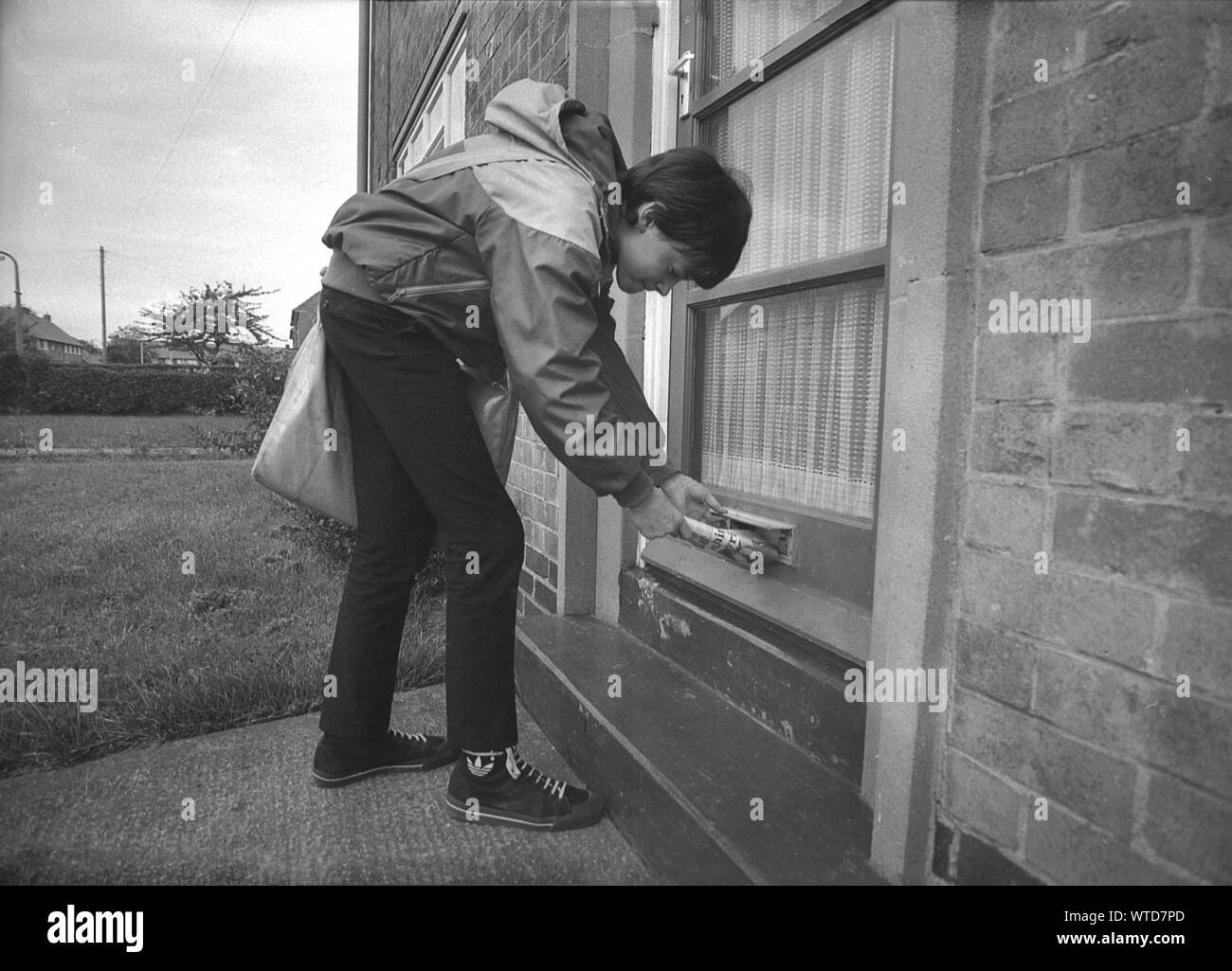 années 1980, historique, un paperboy se penchant vers le bas pour livrer un journal du soir à travers une boîte aux lettres. Après l'école, un garçon de papier avec sacoche, faisant son après-midi ou en début de soirée sur un lotissement était un spectacle courant en Angleterre, au Royaume-Uni. Souvent le premier emploi d'un jeune, il leur a donné leur première expérience de travail et un petit revenu, mais la perte de nombreux journaux imprimés du soir a vu leur nombre diminuer rapidement par rapport aux décennies précédentes. Banque D'Images