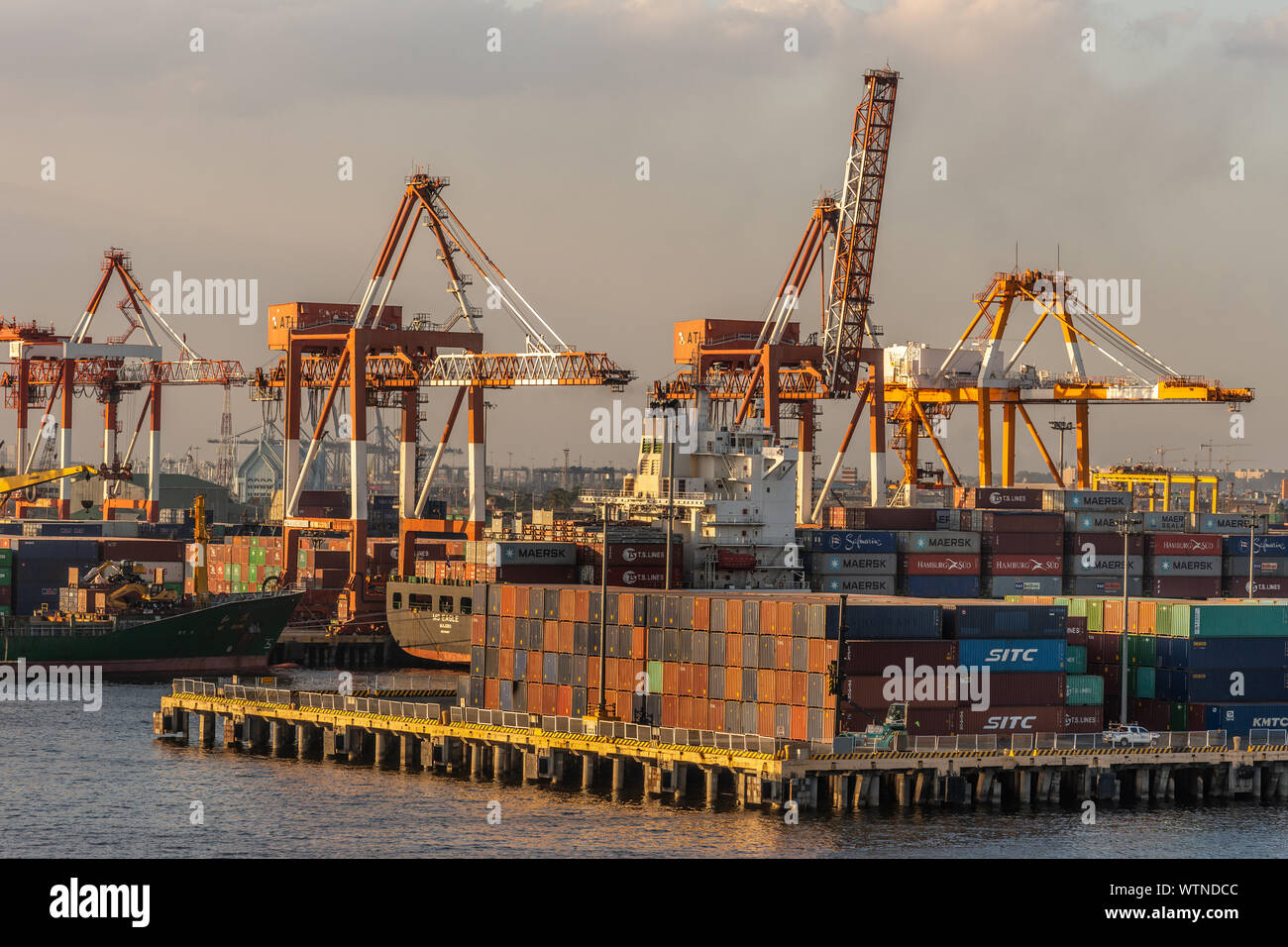 Manille, Philippines - Le 5 mars 2019 : Le port du crépuscule du soir brille sur des piles de conteneurs d'expédition sur la jetée avec les grues et les navires dans le dos. Banque D'Images