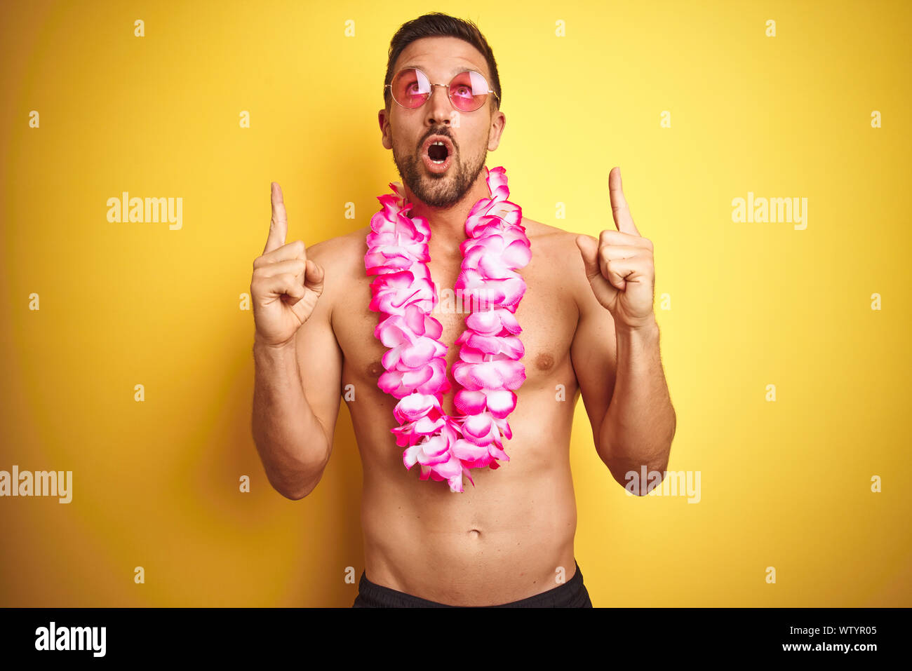 Beau jeune homme torse nu portant des lunettes de soleil et roses Hawaiian  lei sur fond jaune étonné et surpris à la recherche avec les doigts  pointant vers le haut et Photo