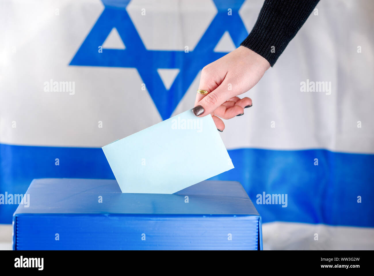 Jeune femme israélienne de mettre un bulletin dans l'urne le jour du scrutin. Close up of hand avec votes blancs sur fond du drapeau d'Israël. Immersive, de l'espace pour le texte. Banque D'Images