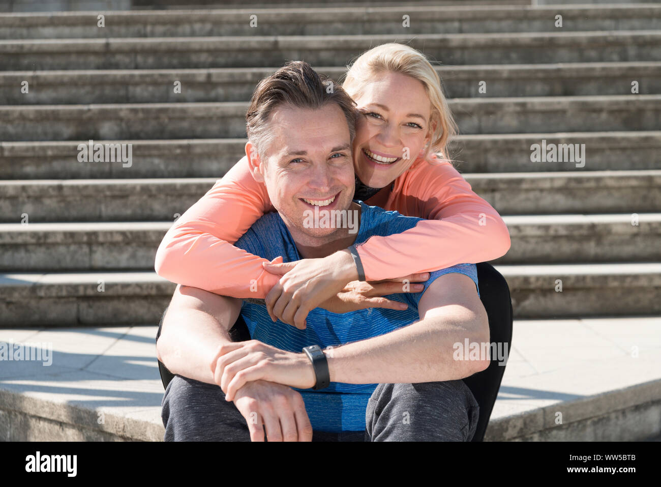 Couple in sportswear sitting on stairs, enlacés, smiling, looking at camera, Banque D'Images