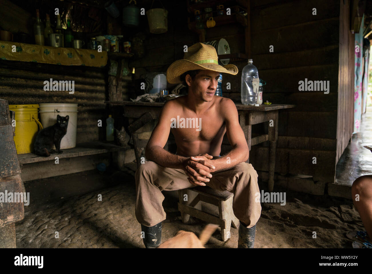 Un jeune agriculteur assis dans la cuisine de sa maison en face de la porte ouverte, des rayons de lumière, lui assis derrière un chat noir Banque D'Images