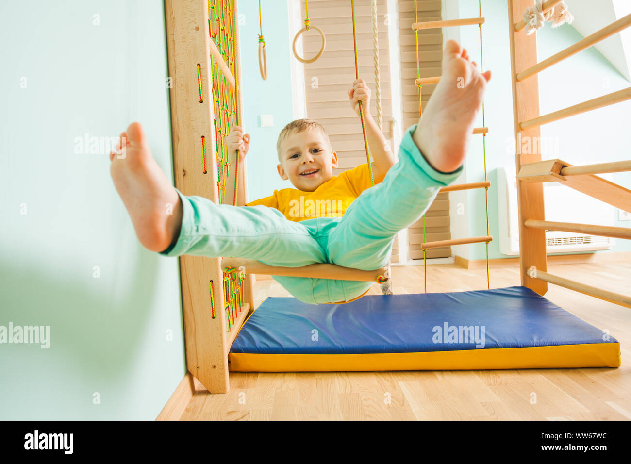 Et l'adorable petit garçon émotif swinging sur une balançoire en bois dans le cadre de kids indoor sport. Petit gars dans un swingigng vêtements colorés sur un sol en bois gy Banque D'Images