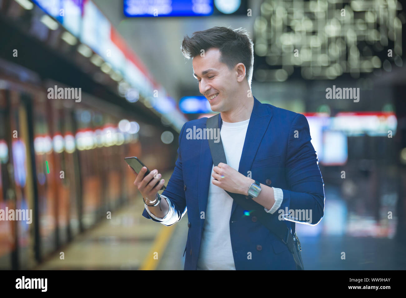 Beau jeune homme élégant en costume avec valise debout sur métro holding smart phone dans la main, et le défilement de texte, sourire et rire. Banque D'Images