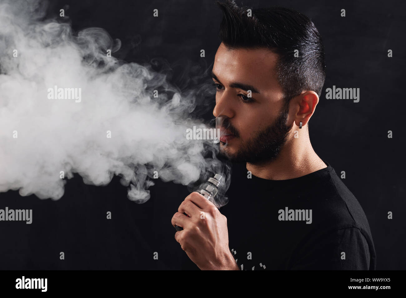 Jeune homme avec barbe vaping cigarette électronique sur fond noir. Banque D'Images
