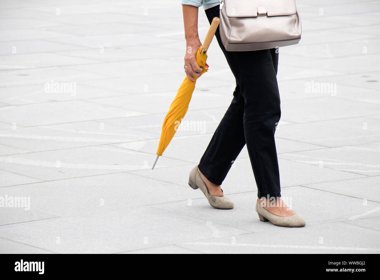 Risque de pluie : une femme marche sur city square pavement exerçant son parapluie jaune pliée , à partir de la taille vers le bas Banque D'Images