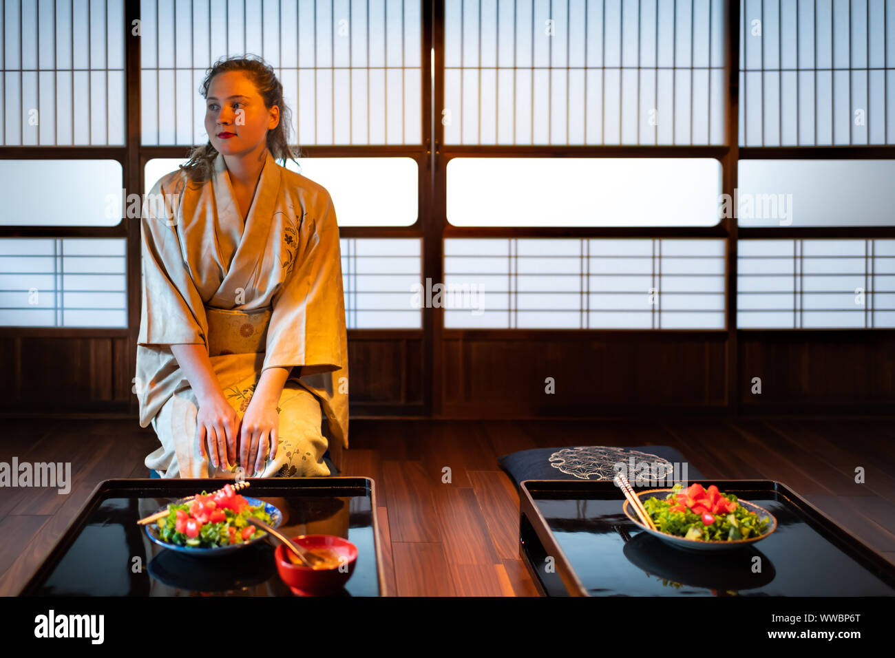Jeune femme en kimono traditionnel japonais assis en seiza chambre ou salle de restaurant ryokan par table avec des assiettes à salade, shoji papier coulissants portes en b Banque D'Images