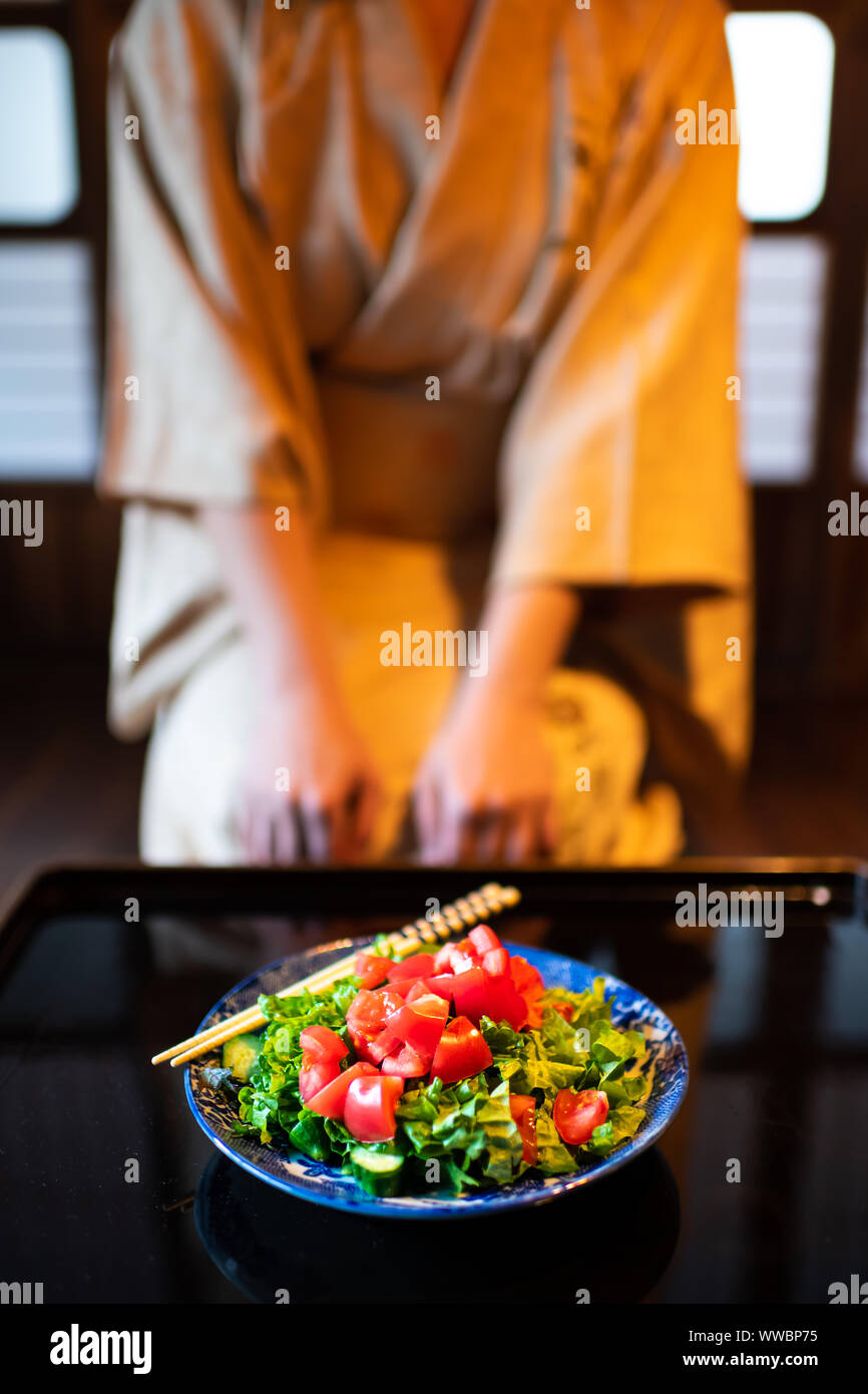 Jeune femme en kimono traditionnel japonais seiza assis à chambre ou salle de restaurant ryokan par table avec salade Assiettes et baguettes, shoji glisser p Banque D'Images