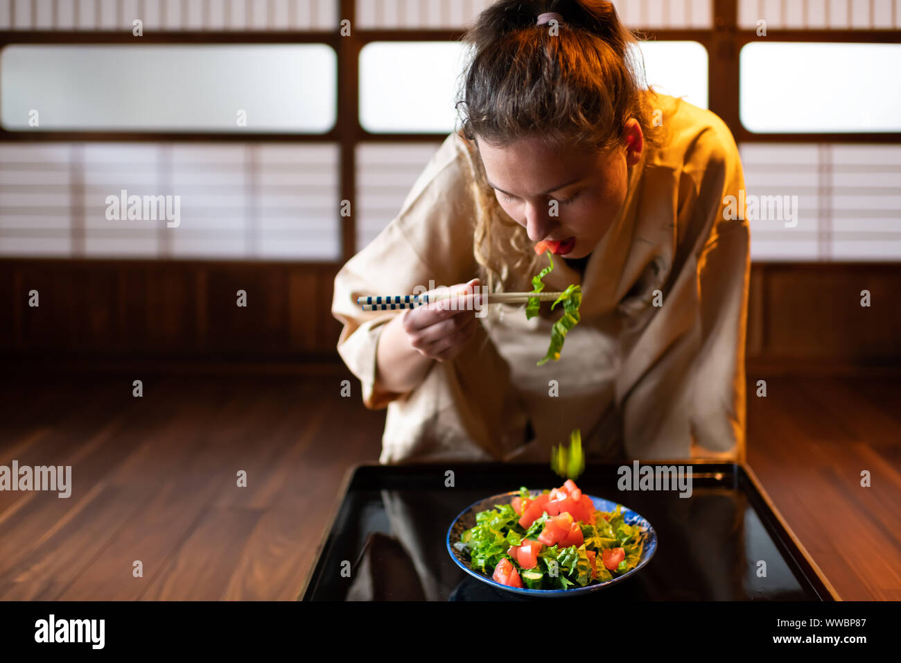 Jeune femme en kimono traditionnel japonais assis à seiza chambre ryokan prix par table de baguettes, mange de la salade par plaque coulissante shoji pa Banque D'Images