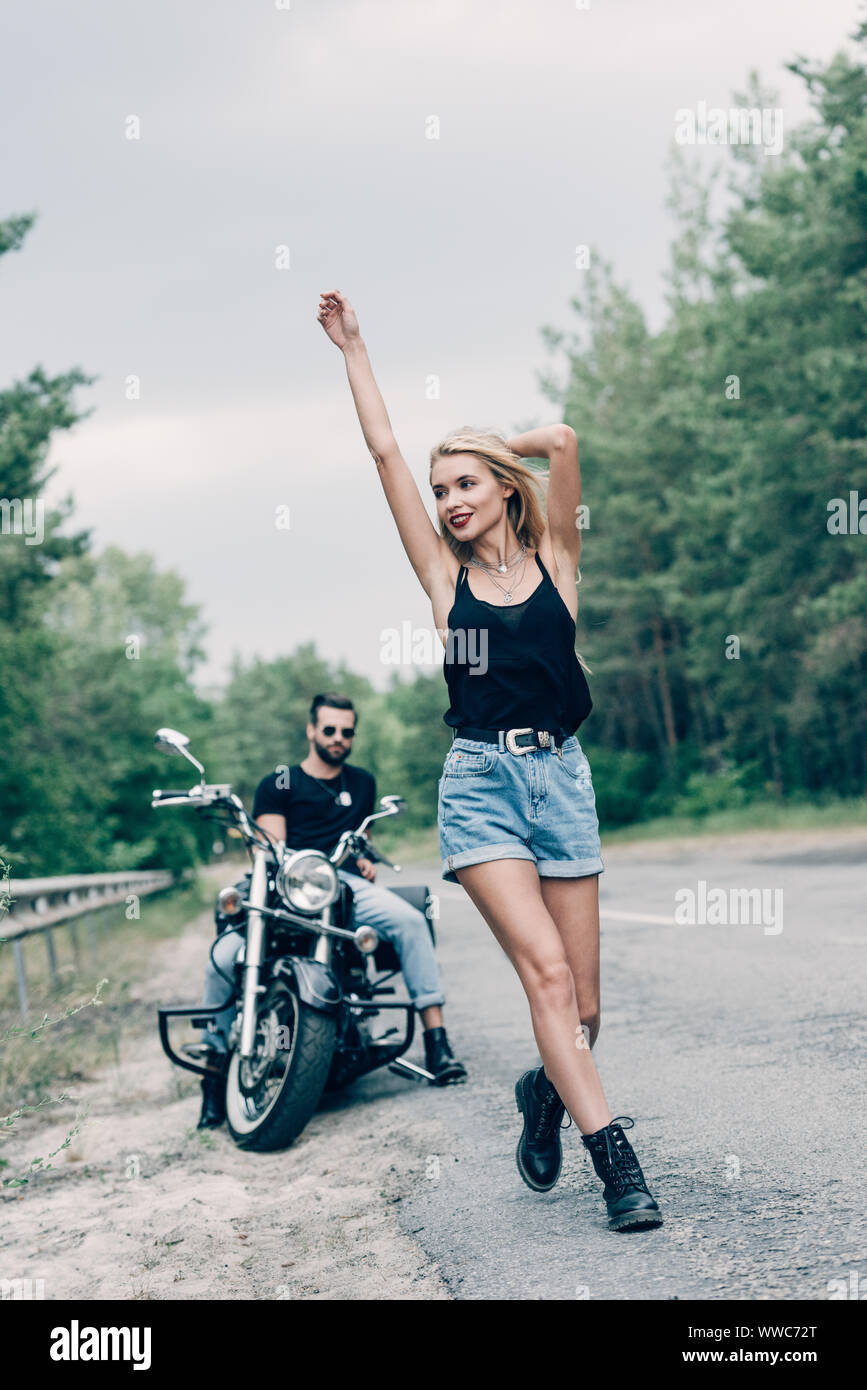 Portrait of happy young woman walking on road avec les mains dans l'air près de petit ami sur moto noir Banque D'Images