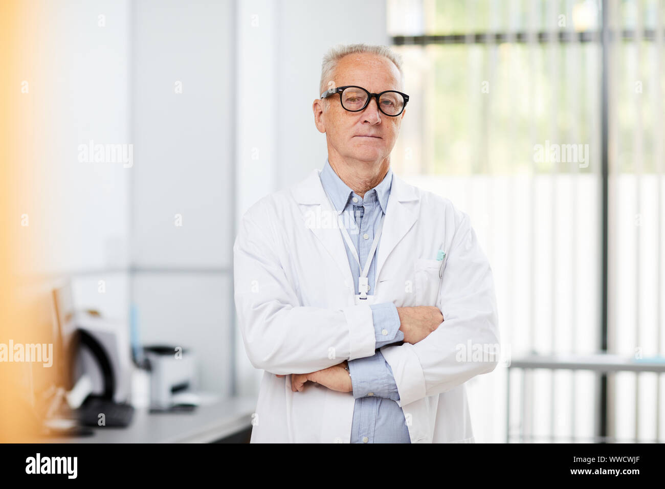 Portrait de friendly senior doctor wearing glasses looking at camera tout en se posant dans office standing with arms crossed, copy space Banque D'Images