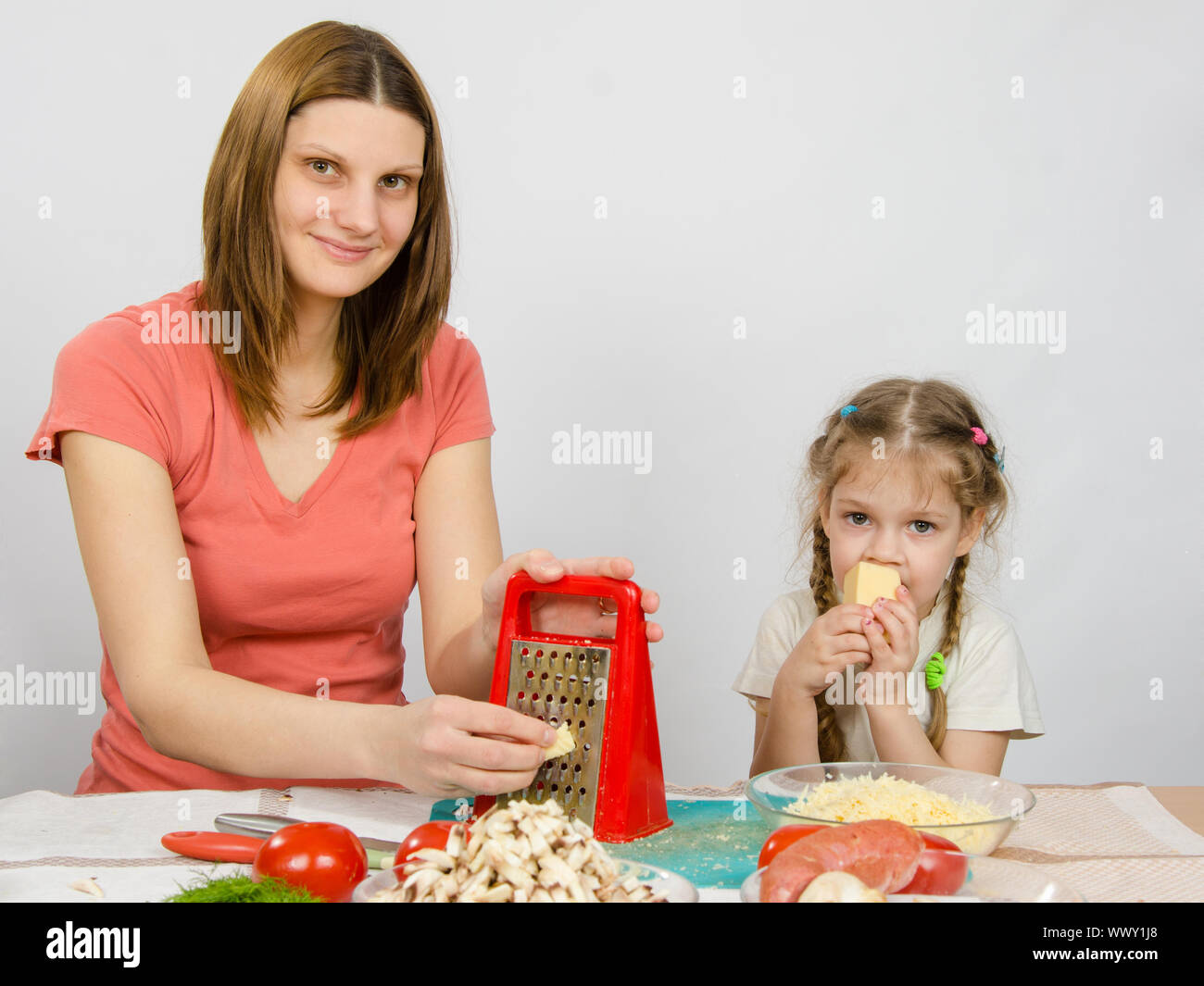 La jeune fille à la table de cuisine râpe à fromage t assis près d'une fille de cinq ans et mange du fromage Banque D'Images